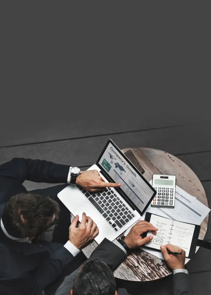 Two business professionals discuss a company's growth report displayed on a laptop, with notes and a calculator on a wooden table.